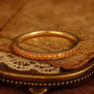 Puede incluir: Un anillo dorado con una banda completamente engastada con pequeñas piedras preciosas de color naranja. El anillo se presenta sobre una superficie marrón texturizada, creando una estética cálida. El diseño del anillo es simple y elegante.