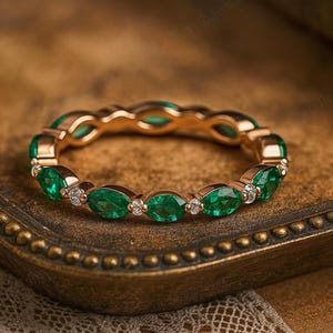 Puede incluir: Un anillo de oro con una serie de esmeraldas de corte ovalado y pequeños diamantes. Las piedras preciosas están engastadas en una banda continua, creando un diseño de eternidad completa. El anillo se muestra en una bandeja de estilo vintage.
