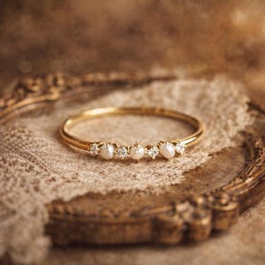 Peut inclure: Une bague en or délicate ornée de petites perles rondes et de pierres précieuses claires. La bague est posée sur un fond aux tons neutres, mettant en valeur son design élégant. La fine bande polie de la bague ajoute à son aspect raffiné.