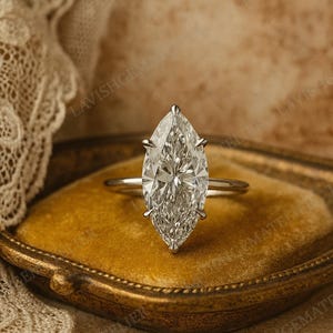 Può includere: Un anello di fidanzamento con diamante taglio marquise e una sottile fascia argentata. Il diamante è incastonato con quattro griffe e brilla intensamente. L'anello è esposto su una superficie di velluto dorato, con il testo "LAVISHGEMATE" visibile sullo sfondo.