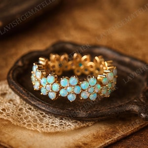 Puede incluir: Un anillo de oro con un diseño floral, adornado con numerosas piedras preciosas opalescentes. La banda del anillo es de oro, y las piedras preciosas exhiben una gama de tonos azules y blancos. El anillo se muestra sobre una superficie oscura y texturizada.