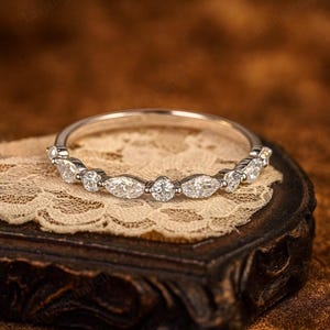 May include: A silver band ring with alternating marquise and round-cut clear gemstones. The ring is set on a vintage wooden box with a lace doily. The gemstones sparkle, reflecting light.