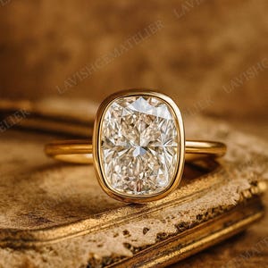 May include: A gold ring with a large, cushion-cut diamond. The diamond is set in a bezel setting and has a brilliant sparkle. The ring is on a textured, golden surface. The words "LAVISHGEMATELIER" are visible.