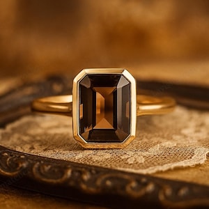 Può includere: Un anello d'oro con una pietra preziosa rettangolare marrone. L'anello è incastonato in una lunetta d'oro e poggia su una superficie simile a un pizzo. Lo sfondo è sfocato e di tonalità calda.