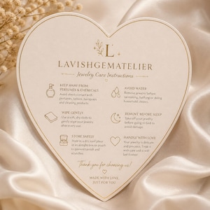 May include: A heart-shaped card with jewelry care instructions. The card is cream-colored with gold text and illustrations. Text includes "Keep Away From Perfumes & Chemicals" and "Handle With Love". The card is on a satin surface.