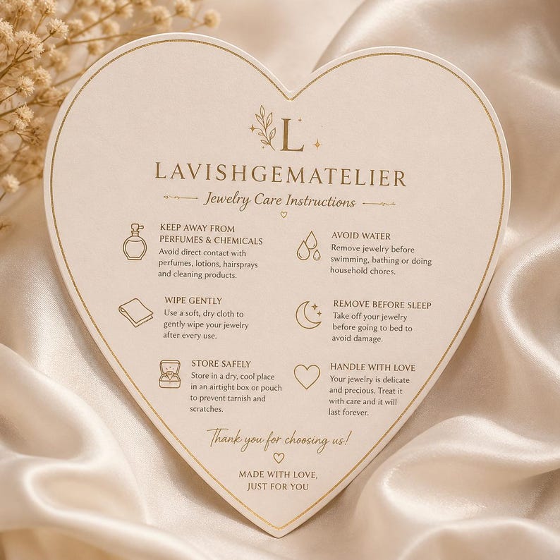 May include: A heart-shaped card with jewelry care instructions. The card is cream-colored with gold text and illustrations. Text includes "Keep Away From Perfumes & Chemicals" and "Handle With Love". The card is on a satin surface.