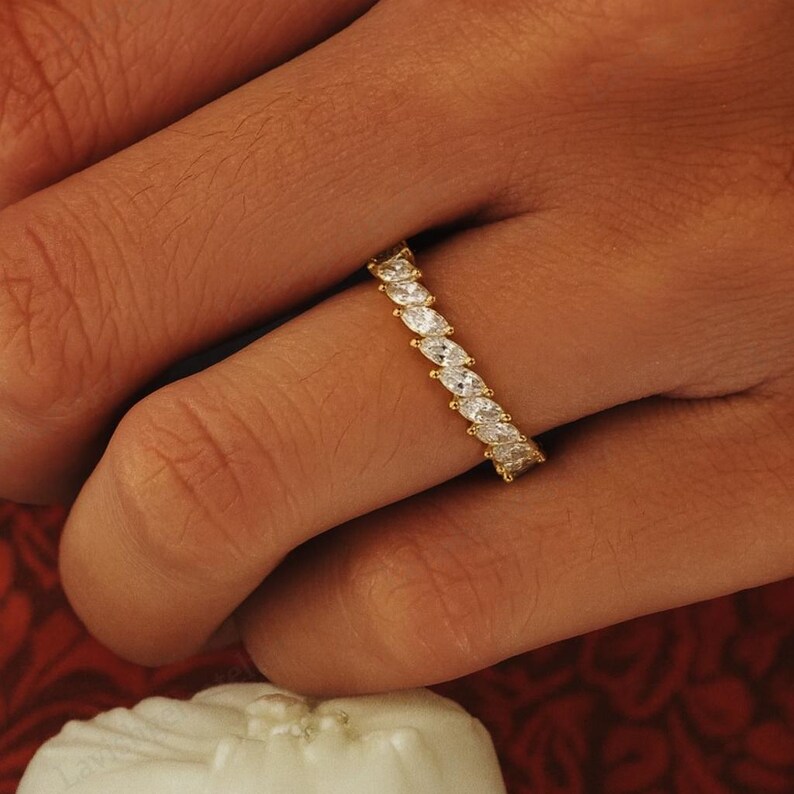 May include: A gold ring with a continuous row of marquise-cut white gemstones. The ring is set on a finger, showcasing the intricate design and sparkle of the gemstones. The ring is a piece of jewelry.