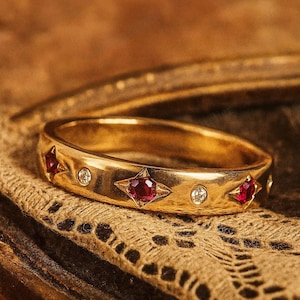 Puede incluir: Un anillo de oro con una banda que presenta estrellas de piedras preciosas rojas y pequeñas gemas claras alternadas. El anillo se coloca sobre un fondo marrón texturizado, resaltando el intrincado diseño y los tonos cálidos de la joya.