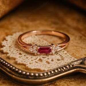 May include: Rose gold ring with a rectangular ruby centre stone and small diamond accents. The ring is set on a delicate band and rests on a lace doily. The ring's design is elegant and suitable for special occasions.