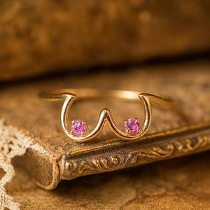 May include: A gold ring with a distinctive design, incorporating two pink gemstones. The ring's form is a stylized representation. The gemstones are round and set within the gold band. The ring is presented on a textured, brown surface.