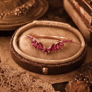 May include: A rose gold ring featuring a curved band set with multiple marquise and round-cut deep red rubies. The stones are arranged in a symmetrical cluster pattern along the center of the band, which also includes small pavé-set red stones.