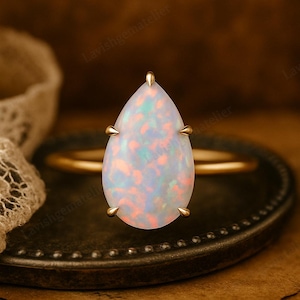 Puede incluir: Un anillo de oro con una piedra de ópalo en forma de pera. El ópalo muestra un juego de colores con tonos rosas, azules y blancos. El anillo tiene una fina banda de oro y cuatro garras doradas que sujetan el ópalo.