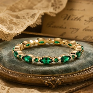 May include: A gold-toned ring featuring an alternating pattern of marquise-cut and round-cut green gemstones set in a continuous band. The metal setting is polished and reflective, highlighting the vibrant emerald-colored stones throughout the circular design.