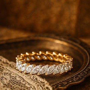 May include: A gold eternity ring with marquise-cut diamonds. The ring is set on a vintage-style brass tray with a lace cloth. The ring's diamonds are clear and sparkling, reflecting light.