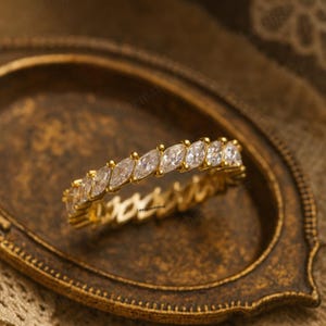 May include: A gold ring with a band of marquise-cut clear stones. The ring is set on a gold band and is displayed on a decorative, antique-style tray. The ring's design is elegant and eye-catching.