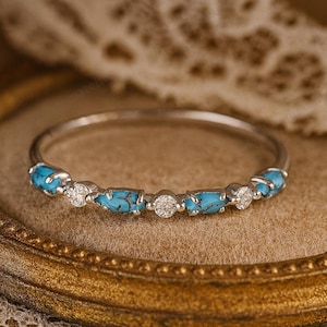 Op de afbeelding: Een zilveren ring met afwisselende turquoise en diamant accenten. De turkooizen stenen zijn helderblauw en de diamanten schitteren. De ring is geplaatst tegen een zachte, neutrale achtergrond.