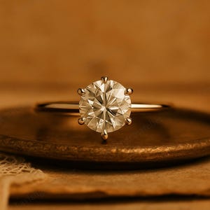 May include: A gold engagement ring featuring a large, round, clear gemstone. The gemstone is held in place by six prongs. The ring is on a brown surface.