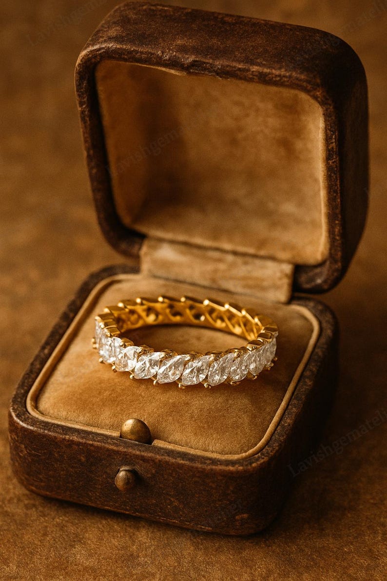 May include: A gold ring with marquise-cut diamonds, displayed in a vintage brown jewelry box. The ring features a continuous row of sparkling gemstones set in a gold band. The box has a velvet interior.