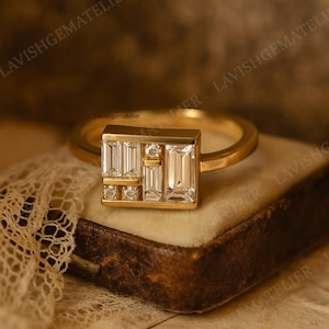 May include: A gold ring with a rectangular face featuring a geometric arrangement of baguette and round-cut diamonds. The ring is set on a vintage-style jewelry box. The text "Lavishgematelier" is visible.