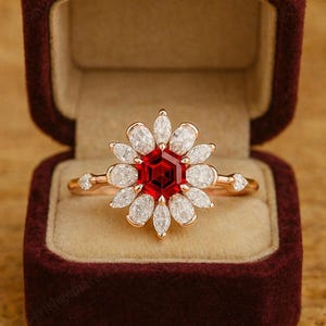 May include: A floral-shaped ring with a red center stone and white petal-shaped stones. The ring is set in rose gold and is displayed in a maroon velvet ring box. The ring's design resembles a flower.