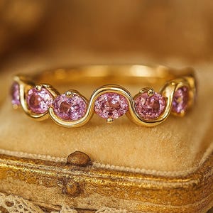 Op de afbeelding: Een gouden ring met een reeks ovale, roze edelstenen. De edelstenen zijn in een golvend patroon langs de band gezet. De ring wordt gepresenteerd op een vintage, goudkleurige sieradendoos met een kanten rand.