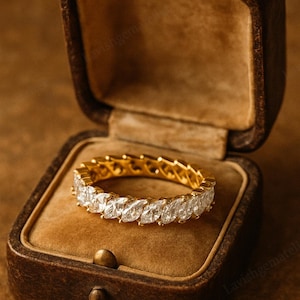 May include: A gold ring with marquise-cut diamonds, displayed in a vintage brown jewelry box. The ring features a continuous row of sparkling gemstones. The box has a velvet interior and a hinged lid, creating a luxurious presentation.