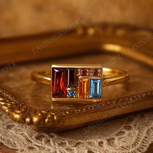 May include: A gold ring with a rectangular face set with colorful gemstones. The ring features a large, rectangular red stone, along with smaller orange, blue, and red stones. The ring is displayed on a gold-colored tray.