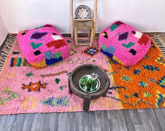 May include: A vibrant pink Moroccan rug with a colorful geometric pattern, complemented by two matching square poufs. A wooden table with a tray and a small decorative chair complete the scene. The rug's design includes orange, blue, and green accents.
