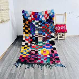 Può includere: Un tappeto rettangolare colorato con un motivo a scacchiera nei toni del giallo, rosa, rosso, blu, verde e nero. Il tappeto ha una frangia lungo il bordo inferiore ed è appoggiato su un pavimento in legno grigio.
