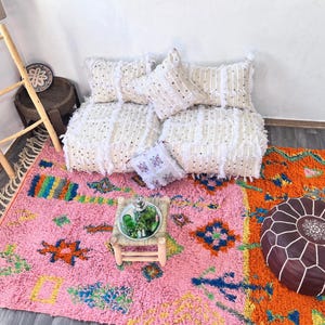 May include: A vibrant, colorful rug with a pink background and geometric patterns in orange, blue, green, and yellow. White fringing edges the rug. White fringed cushions and a brown leather pouf are also visible, creating a cozy, bohemian vibe.