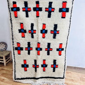 May include: Cream-colored rug with a repeating pattern of black crosses with red and blue accents. The rug has a black border and a fringed edge. The rug is displayed on a wooden floor.