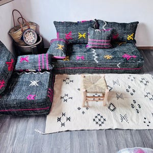 May include: A gray woven sofa and ottoman set with pink, yellow, and white embroidered accents. A matching rug and small wooden chair are in front of the sofa. Woven baskets and a decorative plate are in the background.