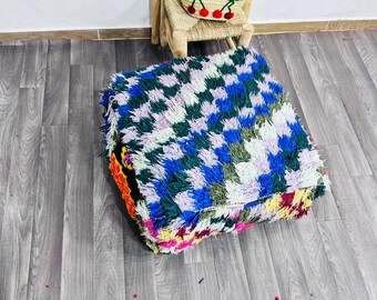 May include: A square, multi-coloured pouf with a chequered pattern in shades of blue, green, white, and purple. The pouf has a shaggy texture and is surrounded by a fringe of various colours. A small woven basket with red handles and cherry embroidery sits on a small wooden chair.