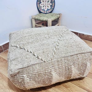 May include: A square, beige woven pouf with a geometric design. The pouf is made of a textured fabric and features a decorative stitched pattern. The pouf is placed on a wooden floor, with a small stool and decorative plate in the background.