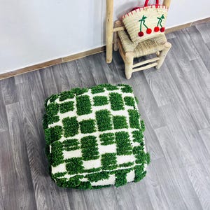 May include: A square, tufted floor cushion with a white and green checkered pattern. A small woven chair holds a straw bag with red cherry accents. A wooden ladder leans against the wall.