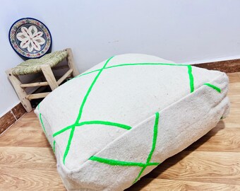Può includere: Un grande pouf quadrato bianco con un design geometrico verde neon. Il cuscino è posto su un pavimento in legno, con un piccolo sgabello in legno e un piatto decorativo sullo sfondo.