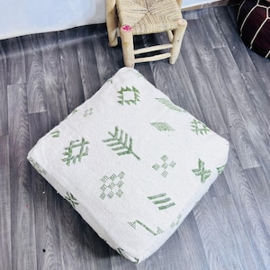 Puede incluir: Cojín de suelo cuadrado blanco con patrones geométricos verdes. El cojín está hecho de una tela texturizada y está colocado sobre un suelo de madera gris. Una pequeña silla de madera con un asiento tejido está en el fondo.