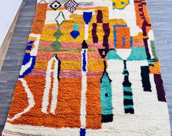 May include: A vibrant, rectangular rug with a colorful abstract design. The rug features a mix of orange, yellow, blue, and white geometric shapes. It has a shaggy texture and a fringe along the edges, adding a bohemian touch to the floor covering.