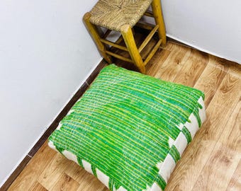May include: A square green and white striped floor cushion. A small, woven stool with a light brown seat and yellow wooden legs is behind the cushion. A decorative plate with a floral design is above the stool.