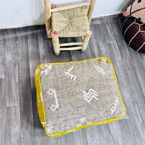 Puede incluir: Un cojín de suelo cuadrado beige con patrones geométricos blancos y un borde amarillo brillante. Una pequeña silla de paja tejida con patas de madera y una escalera de madera están en el fondo. El cojín está sobre un suelo de madera gris.