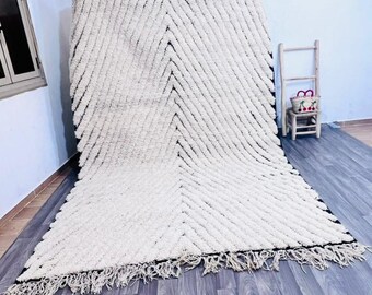 Könnte beinhalten: Ein großer, cremefarbener Teppich mit einem schwarzen geometrischen Muster und Fransen. Der Teppich liegt auf einem grauen Holzboden. Im Hintergrund steht ein kleiner Holzstuhl mit einem Korb. Die Abmessungen des Teppichs sind nicht sichtbar.