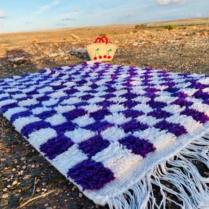 Op de afbeelding: Een wit en paars geruit vloerkleed met franjes en een strooien draagtas met kersendetails. Het vloerkleed ligt buiten op de grond, met een blauwe lucht en een droog landschap op de achtergrond.