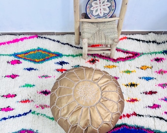 Può includere: Un pouf in pelle dorata con cuciture bianche e un motivo a stella è appoggiato su un colorato tappeto marocchino. Una piccola sedia di legno con seduta intrecciata e un piatto decorativo sono anch'essi nell'immagine.