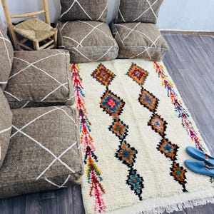 May include: A cream-colored rug with a diamond pattern in orange, blue, and black, and a fringe border. The rug is surrounded by brown cushions with white diamond patterns and a pair of blue shoes.