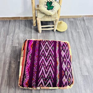 Könnte beinhalten: Quadratisches Bodenkissen mit geometrischem Muster in Lila, Rosa und Orange. Das Kissen hat einen cremefarbenen Rand und liegt auf einem Holzboden. Ein kleiner Strohstuhl und eine Tasche mit Kaktusmotiv befinden sich im Hintergrund.