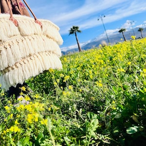 Könnte beinhalten: Eine cremefarbene Makramee-Tragetasche mit langen Fransen wird vor dem Hintergrund eines Feldes mit gelben Blumen, Palmen und blauem Himmel gehalten. Die Tasche hat braune Ledergriffe und Akzente.