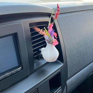 Peut inclure: Un vase blanc imprimé en 3D pour aération de voiture contenant des fleurs artificielles colorées. Le vase, en forme de petit pichet, est inséré dans une bouche d'aération de voiture. Les fleurs comprennent des fleurs roses, violettes et rouges.