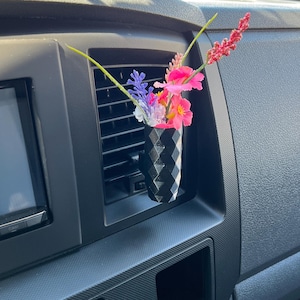 Peut inclure: Un vase noir géométrique pour aérateur de voiture contenant des fleurs artificielles colorées. Le vase est inséré dans une bouche d'aération de voiture, avec des fleurs roses, violettes et jaunes pour décorer l'intérieur du véhicule.