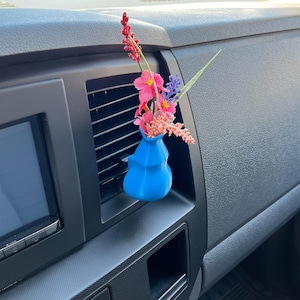May include: A bright blue, 3D-printed car vent vase holding a small bouquet of colorful artificial flowers. The flowers include pink, red, and purple blooms, adding a touch of color to the car's interior.
