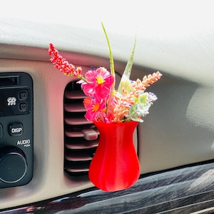 Peut inclure: Un vase rouge vif imprimé en 3D pour aérateur de voiture, rempli de fleurs artificielles colorées. Le vase est conçu pour se fixer sur une grille d'aération de voiture, ajoutant une touche décorative. Les fleurs présentent des accents roses, rouges et verts.